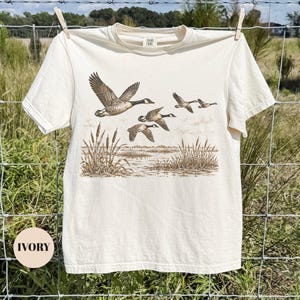 Peut inclure: Un t-shirt &agrave; manches courtes ivoire avec une impression graphique marron style encre repr&eacute;sentant un vol d'oies au-dessus d'un marais avec des roseaux. Le t-shirt a un col rond et une &eacute;tiquette au col int&eacute;rieur indiquant Comfort Colors.