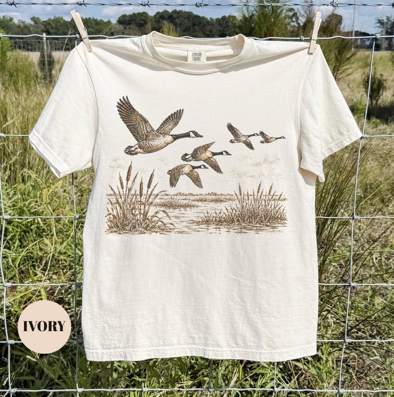 Peut inclure: Un t-shirt &agrave; manches courtes ivoire avec une impression graphique marron style encre repr&eacute;sentant un vol d'oies au-dessus d'un marais avec des roseaux. Le t-shirt a un col rond et une &eacute;tiquette au col int&eacute;rieur indiquant Comfort Colors.