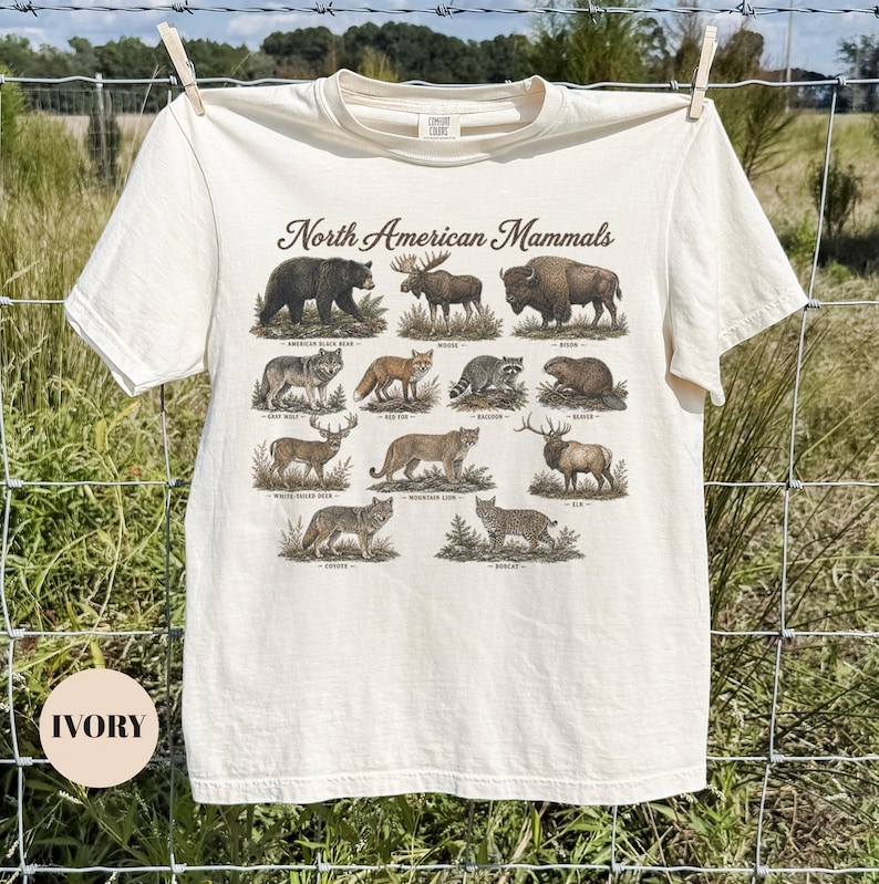 May include: An ivory short-sleeve t-shirt featuring a graphic print titled North American Mammals. The design displays illustrations of a black bear, moose, bison, wolf, fox, raccoon, beaver, deer, mountain lion, elk, coyote, and bobcat with labels.