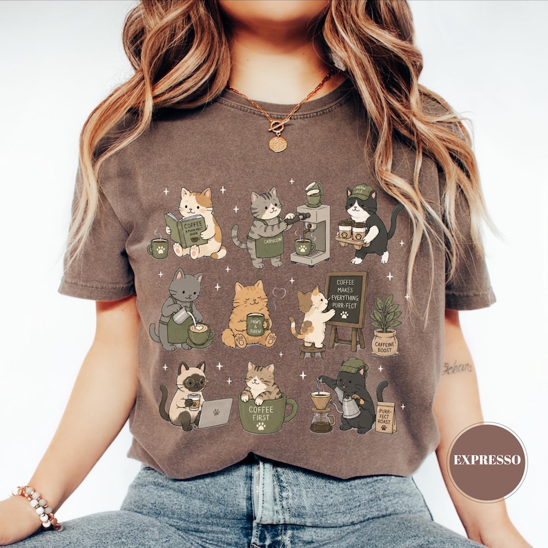 May include: Brown t-shirt featuring a whimsical design of various cats engaged in coffee-related activities. The cats are depicted reading, brewing, and enjoying coffee, with text that reads "Coffee Makes Everything Purr-fect."