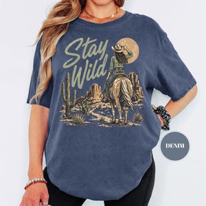 May include: Navy blue t-shirt featuring a desert landscape design. The graphic depicts a cowgirl riding a horse, with cacti and the words "Stay Wild". The shirt has short sleeves and a loose fit.