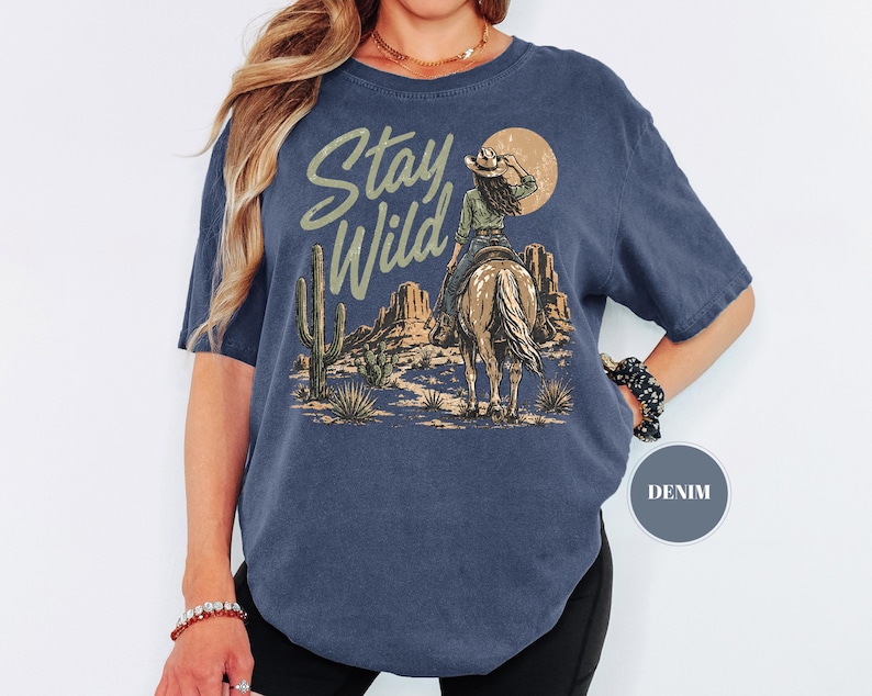 May include: Navy blue t-shirt featuring a desert landscape design. The graphic depicts a cowgirl riding a horse, with cacti and the words "Stay Wild". The shirt has short sleeves and a loose fit.