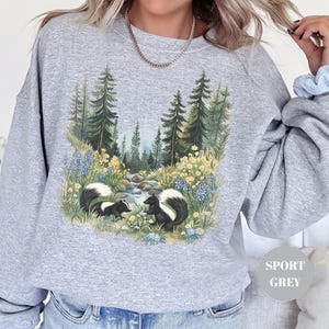 May include: Sport grey sweatshirt featuring a nature scene with two skunks near a stream, surrounded by trees and wildflowers. The graphic is detailed with shades of green, blue, and yellow. The sweatshirt has long sleeves and a relaxed fit.
