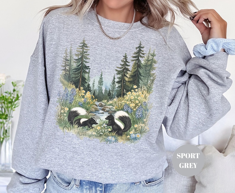 May include: Sport grey sweatshirt featuring a nature scene with two skunks near a stream, surrounded by trees and wildflowers. The graphic is detailed with shades of green, blue, and yellow. The sweatshirt has long sleeves and a relaxed fit.