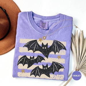 May include: A violet short-sleeve t-shirt with a crew neckline featuring a graphic of three black bats and small gold stars over horizontal beige stripes. The shirt has a Comfort Colors brand label on the inside neck with size S, P, CH.