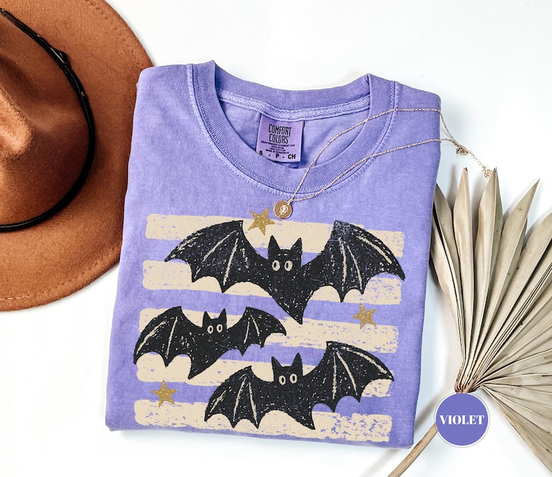 May include: A violet short-sleeve t-shirt with a crew neckline featuring a graphic of three black bats and small gold stars over horizontal beige stripes. The shirt has a Comfort Colors brand label on the inside neck with size S, P, CH.
