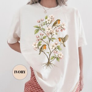 May include: Ivory-colored t-shirt featuring a detailed illustration of a flowering tree with three robins. The birds have orange breasts and are perched among white blossoms and green leaves. The shirt has a relaxed fit.
