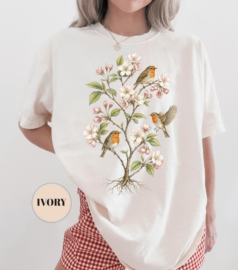 May include: Ivory-colored t-shirt featuring a detailed illustration of a flowering tree with three robins. The birds have orange breasts and are perched among white blossoms and green leaves. The shirt has a relaxed fit.