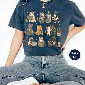 May include: Navy blue t-shirt featuring a whimsical design of cats engaged in coffee-related activities. The cats are depicted brewing coffee, reading, and enjoying their coffee. The shirt also has the text "Coffee Makes Everything Purr-fect".
