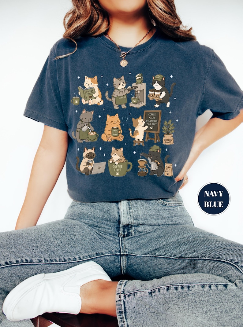 May include: Navy blue t-shirt featuring a whimsical design of cats engaged in coffee-related activities. The cats are depicted brewing coffee, reading, and enjoying their coffee. The shirt also has the text "Coffee Makes Everything Purr-fect".