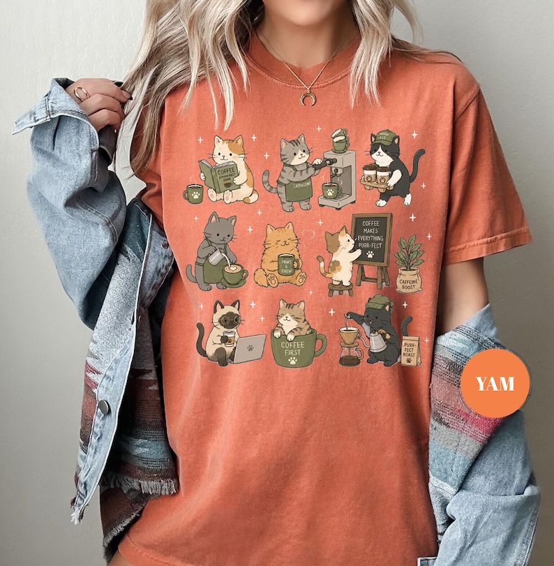May include: Orange t-shirt featuring a whimsical illustration of various cats engaged in coffee-related activities. Cats are reading, brewing, and enjoying coffee. The phrase "Coffee Makes Everything Purr-fect" is on a small chalkboard. The shirt has a relaxed fit.