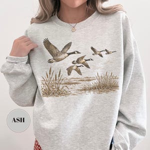 Peut inclure: Un sweat-shirt &agrave; col rond gris cendr&eacute; arborant une impression graphique de quatre oies en vol au-dessus d'un paysage mar&eacute;cageux avec de grands roseaux. Le motif est rendu dans des tons de brun et de beige. Une &eacute;tiquette affiche le texte ASH.