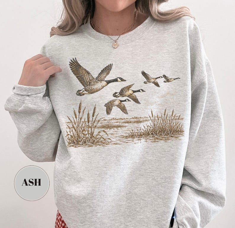 Peut inclure: Un sweat-shirt &agrave; col rond gris cendr&eacute; arborant une impression graphique de quatre oies en vol au-dessus d'un paysage mar&eacute;cageux avec de grands roseaux. Le motif est rendu dans des tons de brun et de beige. Une &eacute;tiquette affiche le texte ASH.