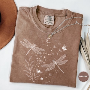 May include: A brown Comfort Colors t-shirt with a white dragonfly and floral design. The shirt has a crew neck and short sleeves. A silver necklace with a moon pendant is on the shirt.