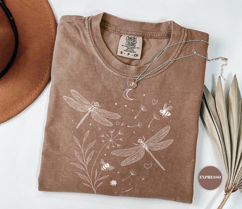 May include: A brown Comfort Colors t-shirt with a white dragonfly and floral design. The shirt has a crew neck and short sleeves. A silver necklace with a moon pendant is on the shirt.