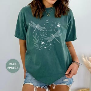 May include: Blue spruce t-shirt with a white dragonfly and bee graphic. The design includes a crescent moon, hearts, and floral elements. The shirt is a short-sleeved, crew neck style.