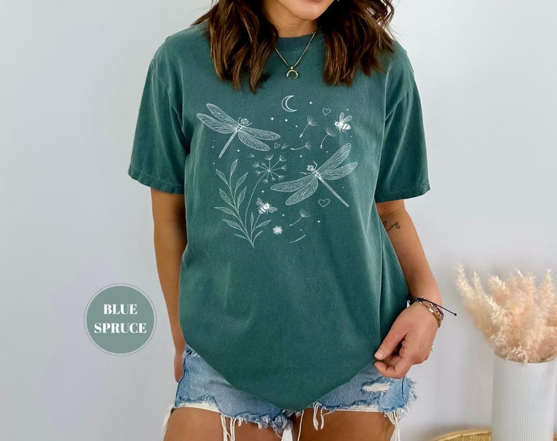 May include: Blue spruce t-shirt with a white dragonfly and bee graphic. The design includes a crescent moon, hearts, and floral elements. The shirt is a short-sleeved, crew neck style.