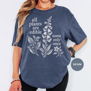 以下が含まれることがあります： デニムブルーの半袖Tシャツで、ジギタリスとベリーの白い植物画が描かれています。白い文字で「all plants are edible some only once」とプリントされています。クルーネックでゆったりとしたフィット感のTシャツです。
