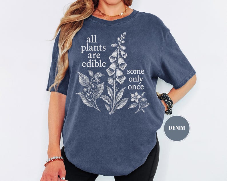 以下が含まれることがあります： デニムブルーの半袖Tシャツで、ジギタリスとベリーの白い植物画が描かれています。白い文字で「all plants are edible some only once」とプリントされています。クルーネックでゆったりとしたフィット感のTシャツです。