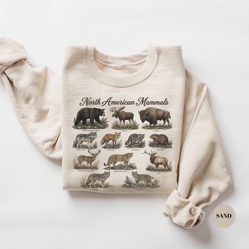 May include: A sand-colored crewneck sweatshirt featuring a graphic print titled North American Mammals. The design displays detailed illustrations of various animals including a black bear, moose, bison, wolf, fox, raccoon, beaver, deer, cougar, elk, coyote, and bobcat, each with its name printed below.