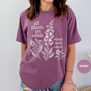 以下が含まれることがあります： ベリー色の半袖Tシャツ。前面には白い植物のイラストと「all plants are edible some only once」というテキストがプリントされています。クルーネックで、ゆったりとしたカジュアルなフィット感のTシャツです。