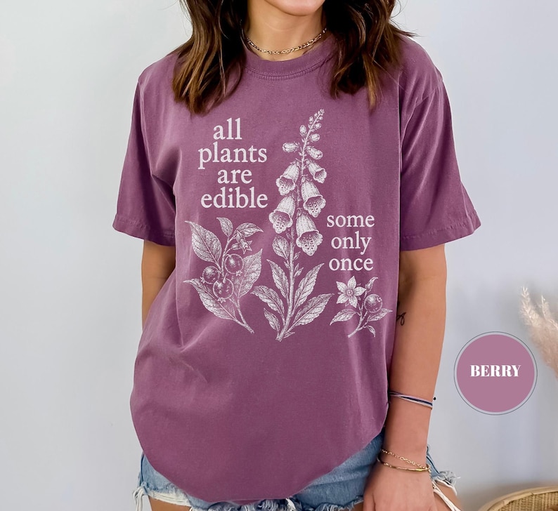 以下が含まれることがあります： ベリー色の半袖Tシャツ。前面には白い植物のイラストと「all plants are edible some only once」というテキストがプリントされています。クルーネックで、ゆったりとしたカジュアルなフィット感のTシャツです。