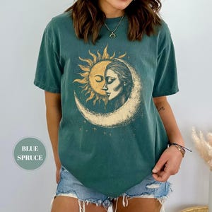May include: Blue spruce t-shirt featuring a sun and moon graphic. The design shows a sun embracing a woman's face within a crescent moon. The shirt has short sleeves and a relaxed fit, perfect for casual wear.
