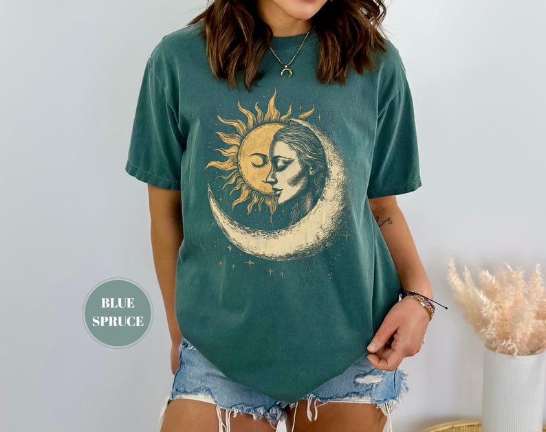 May include: Blue spruce t-shirt featuring a sun and moon graphic. The design shows a sun embracing a woman's face within a crescent moon. The shirt has short sleeves and a relaxed fit, perfect for casual wear.