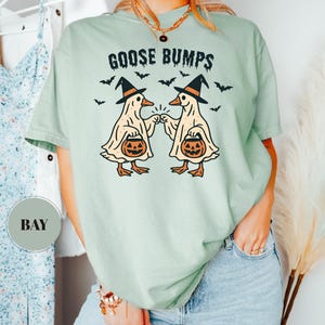 May include: A light green t-shirt with the words "GOOSE BUMPS" above a graphic of two cartoon geese wearing witch hats and holding jack-o'-lanterns. Black bats are scattered around the geese. The shirt is a comfortable, casual style.