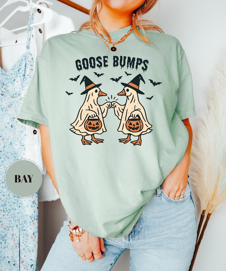 May include: A light green t-shirt with the words "GOOSE BUMPS" above a graphic of two cartoon geese wearing witch hats and holding jack-o'-lanterns. Black bats are scattered around the geese. The shirt is a comfortable, casual style.