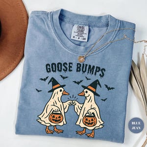 May include: A blue jean Comfort Colors t-shirt with the words "GOOSE BUMPS" in black. The shirt features a graphic of two cartoon geese dressed as ghosts, holding jack-o'-lanterns, and wearing witch hats. Bats are scattered around the geese.