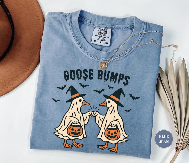 May include: A blue jean Comfort Colors t-shirt with the words "GOOSE BUMPS" in black. The shirt features a graphic of two cartoon geese dressed as ghosts, holding jack-o'-lanterns, and wearing witch hats. Bats are scattered around the geese.