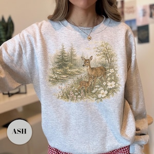 May include: Ash-colored sweatshirt featuring a detailed illustration of two deer in a forest scene. The design includes a doe and a fawn near a stream, surrounded by trees and flowers. The sweatshirt has a classic crew neck.