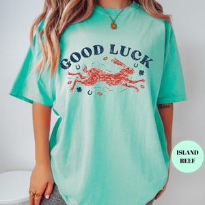 K&ouml;nnte beinhalten: T&uuml;rkisfarbenes T-Shirt mit dem Schriftzug "GOOD LUCK" in Marineblau &uuml;ber einer Illustration eines roten Hasen. Das Design beinhaltet Hufeisen und Kleebl&auml;tter. Das Shirt hat kurze &Auml;rmel und eine lockere Passform.