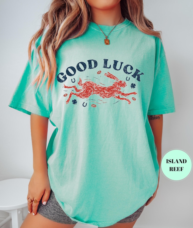 K&ouml;nnte beinhalten: T&uuml;rkisfarbenes T-Shirt mit dem Schriftzug "GOOD LUCK" in Marineblau &uuml;ber einer Illustration eines roten Hasen. Das Design beinhaltet Hufeisen und Kleebl&auml;tter. Das Shirt hat kurze &Auml;rmel und eine lockere Passform.