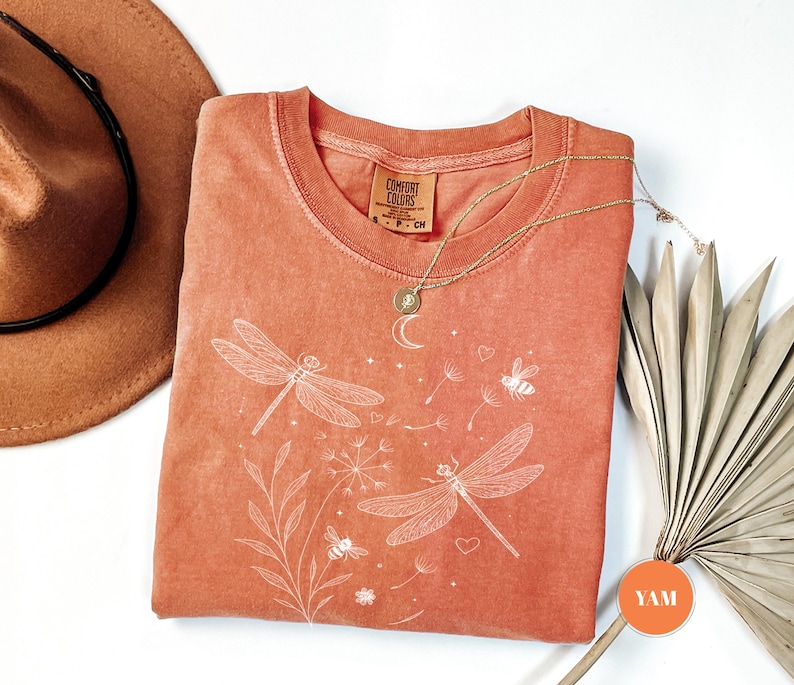 May include: Coral-colored Comfort Colors t-shirt with a crew neck, featuring a white dragonfly and floral design. A gold necklace with a pendant rests on the shirt. A brown hat and dried palm leaves are in the background.