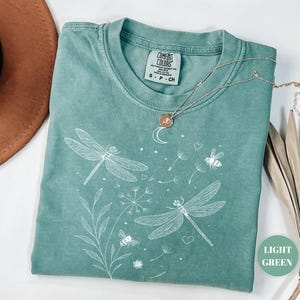 May include: Light green t-shirt with a dragonfly and bee graphic. The shirt features a crew neck and short sleeves. A silver necklace with a pendant is draped across the shirt. The shirt is folded on a white surface.