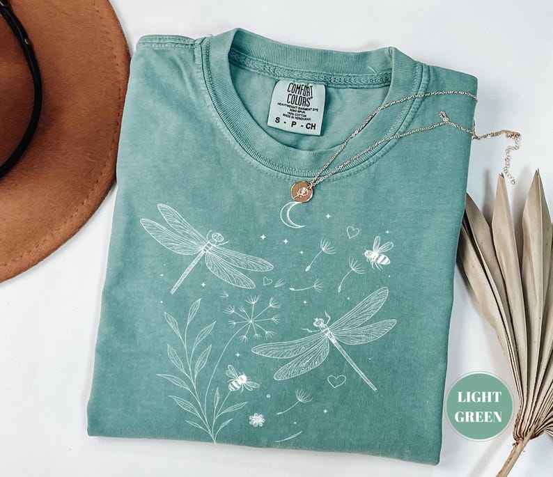 May include: Light green t-shirt with a dragonfly and bee graphic. The shirt features a crew neck and short sleeves. A silver necklace with a pendant is draped across the shirt. The shirt is folded on a white surface.