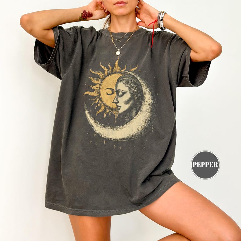 May include: Dark gray t-shirt featuring a sun and moon graphic with a woman's face. The design is in shades of gold and white. The shirt has short sleeves and a relaxed fit.