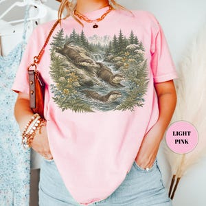 Puede incluir: Una camiseta rosa claro con un estampado gr&aacute;fico detallado de tres nutrias nadando y jugando en un arroyo forestal rodeado de pinos, helechos y flores silvestres. El dise&ntilde;o est&aacute; realizado en tonos tierra apagados que contrastan con la tela rosa.