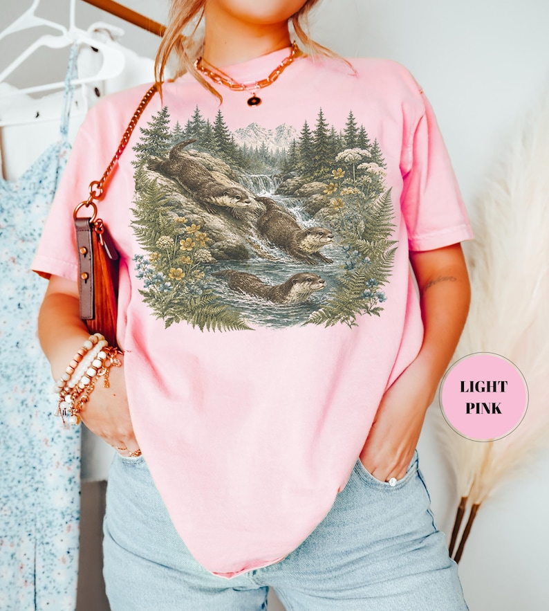 Puede incluir: Una camiseta rosa claro con un estampado gr&aacute;fico detallado de tres nutrias nadando y jugando en un arroyo forestal rodeado de pinos, helechos y flores silvestres. El dise&ntilde;o est&aacute; realizado en tonos tierra apagados que contrastan con la tela rosa.