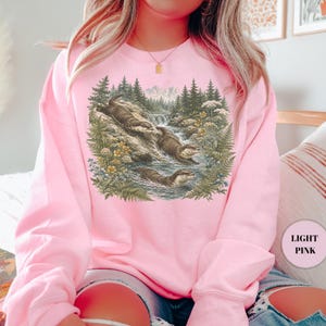 Puede incluir: Una sudadera de cuello redondo en color rosa claro con un estampado gr&aacute;fico detallado de tres nutrias en un arroyo forestal. La ilustraci&oacute;n muestra a las nutrias nadando y descansando sobre rocas entre pinos, helechos y flores silvestres.