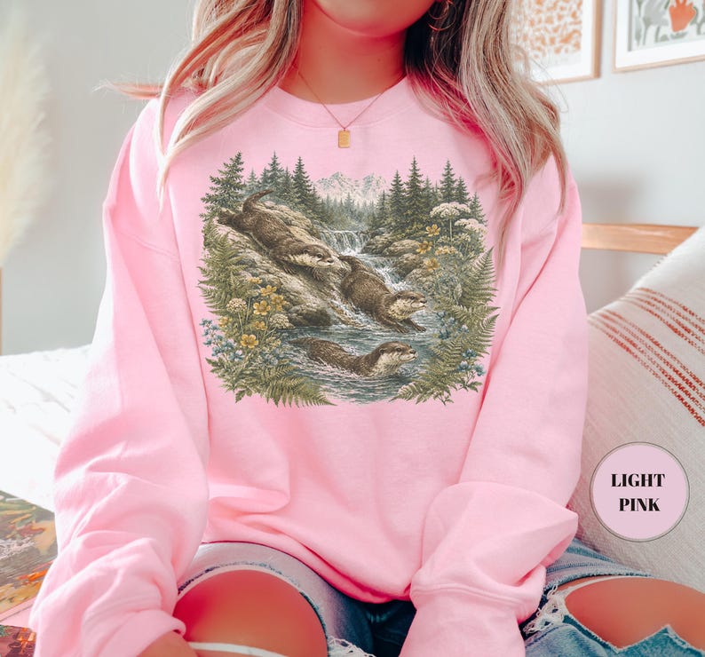 Puede incluir: Una sudadera de cuello redondo en color rosa claro con un estampado gr&aacute;fico detallado de tres nutrias en un arroyo forestal. La ilustraci&oacute;n muestra a las nutrias nadando y descansando sobre rocas entre pinos, helechos y flores silvestres.