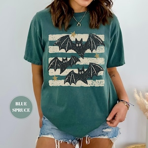 May include: A blue spruce colored short-sleeve t-shirt featuring a graphic print of three black bats flying over horizontal distressed white stripes with small gold stars scattered throughout the design. The shirt has a crew neckline and a relaxed fit.