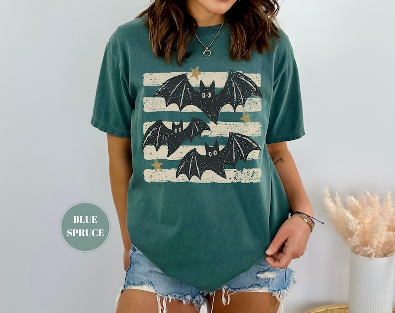 May include: A blue spruce colored short-sleeve t-shirt featuring a graphic print of three black bats flying over horizontal distressed white stripes with small gold stars scattered throughout the design. The shirt has a crew neckline and a relaxed fit.