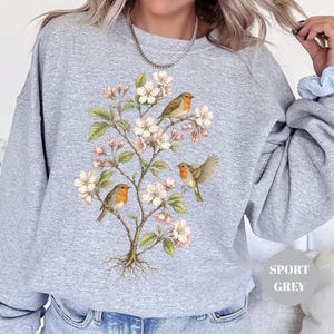 May include: A light gray sweatshirt featuring a botanical illustration of a flowering tree with three robins. The birds have orange breasts and are perched on branches with white blossoms and green leaves. The sweatshirt has the words "Sport Grey" printed on it.