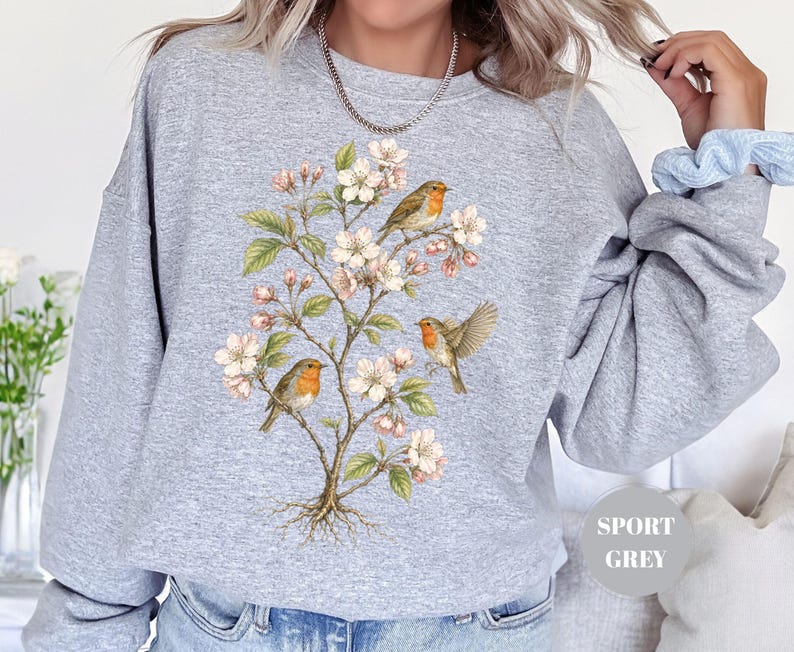 May include: A light gray sweatshirt featuring a botanical illustration of a flowering tree with three robins. The birds have orange breasts and are perched on branches with white blossoms and green leaves. The sweatshirt has the words "Sport Grey" printed on it.