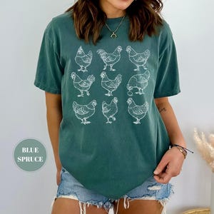 May include: A blue spruce t-shirt featuring a white graphic of nine chickens. The short-sleeved shirt is made of a soft material and is a relaxed fit. The text "BLUE SPRUCE" is in a circle on the bottom left of the image.