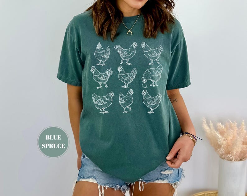 May include: A blue spruce t-shirt featuring a white graphic of nine chickens. The short-sleeved shirt is made of a soft material and is a relaxed fit. The text "BLUE SPRUCE" is in a circle on the bottom left of the image.