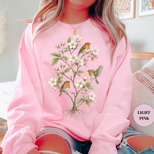 May include: Light pink sweatshirt featuring a detailed illustration of a flowering tree with three birds perched on its branches. The birds have orange breasts and brown wings. The tree has white blossoms and green leaves.
